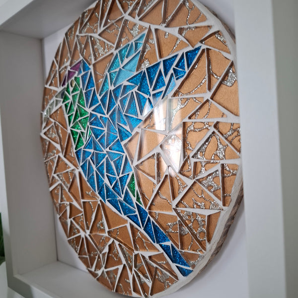 A circular mosaic wall art piece mounted inside a white recessed shelf. The design features a hummingbird in blue, green, and purple tiles separated by white grout lines, with a glossy reflective center.