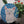 Circular mosaic wall art featuring a stylized cat design. Crafted with glittery white, silver and pink glass pieces on a sparkly blue background, set on rustic wood. Displayed on a wall in a room with plants to show how beautiful the piece is in a home. 