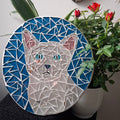 Circular mosaic wall art featuring a stylized cat design. Crafted with glittery white, silver and pink glass pieces on a sparkly blue background, set on rustic wood. Displayed on a wall in a room with plants to show how beautiful the piece is in a home. 