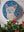 Circular mosaic wall art featuring a stylized cat design. Crafted with glittery white, silver and pink glass pieces on a sparkly blue background, set on rustic wood. Displayed on a wall in a room with plants.