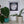 A framed circular mosaic wall art piece featuring a dragonfly with blue, white, and pink geometric tiles, displayed on a shelf above green potted plants.