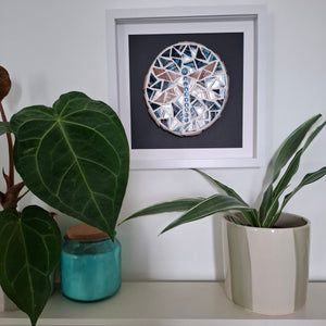 A framed circular mosaic wall art piece featuring a dragonfly with blue, white, and pink geometric tiles, displayed on a shelf above green potted plants.