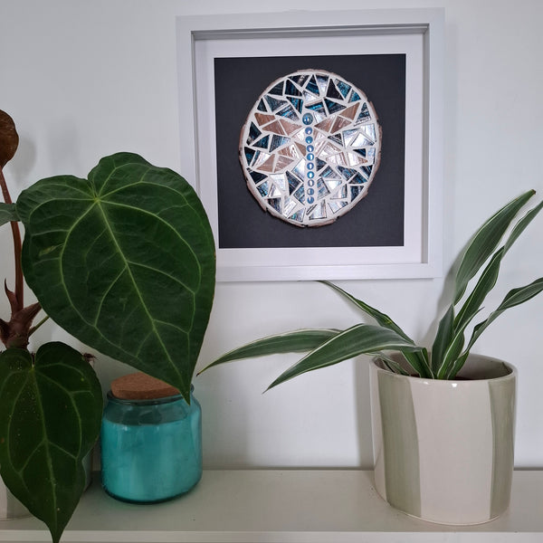 A framed circular mosaic wall art piece featuring a dragonfly with blue, white, and pink geometric tiles, displayed on a shelf above green potted plants.