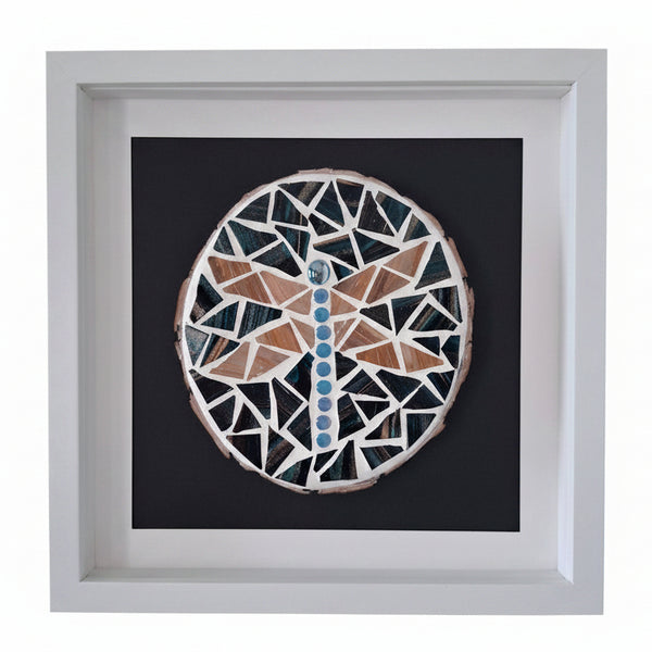 A square framed wall art piece featuring a circular mosaic dragonfly design in pink, white, and blue on a black background, mounted in a white frame against a light gray wall.