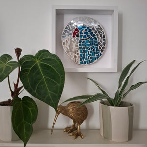 A square white shadow box wall decor piece featuring a colorful mosaic parrot, styled on a shelf with potted plants and a gold kiwi bird figurine.
