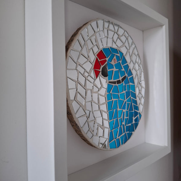 A circular mosaic wall art piece featuring a stylized bird design in blue, red, black, and white, mounted inside a white recessed wall frame.