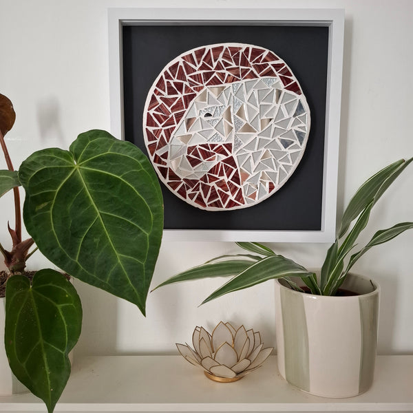 A framed mosaic elephant wall art piece hangs on a white wall above a shelf with potted plants and a small decorative lotus holder. The artwork features a geometric mosaic design in silver, terracotta red, and dark brown tones on a black background, enclosed in a white frame.