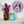 A circular mosaic wall art piece featuring a stylized bat in shades of purple, blue, black, and orange, mounted on a white wall above a shelf with an amber glass vase of purple flowers and a turquoise glass jar.