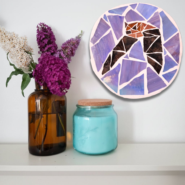 A circular mosaic wall art piece featuring a stylized bat in shades of purple, blue, black, and orange, mounted on a white wall above a shelf with an amber glass vase of purple flowers and a turquoise glass jar.