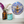 A round mosaic wall art piece featuring a stylized bee in blue, silver, black, and gold tones, displayed on a white shelf next to a brown glass vase with flowers and a turquoise glass jar.