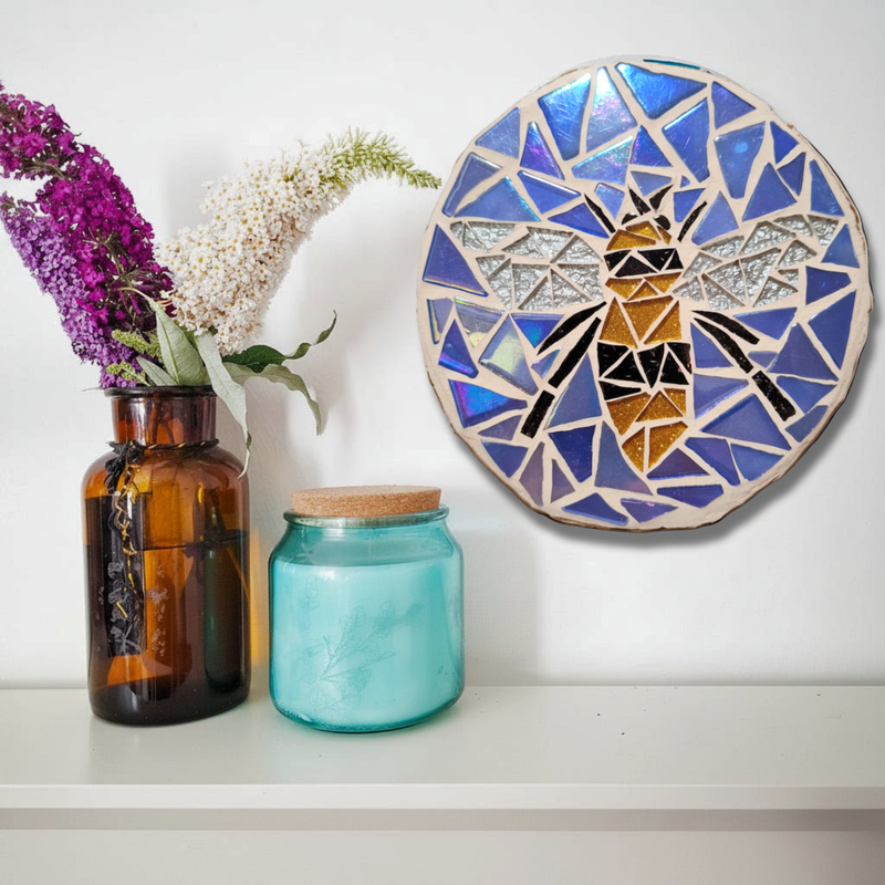 A round mosaic wall art piece featuring a stylized bee in blue, silver, black, and gold tones, displayed on a white shelf next to a brown glass vase with flowers and a turquoise glass jar.