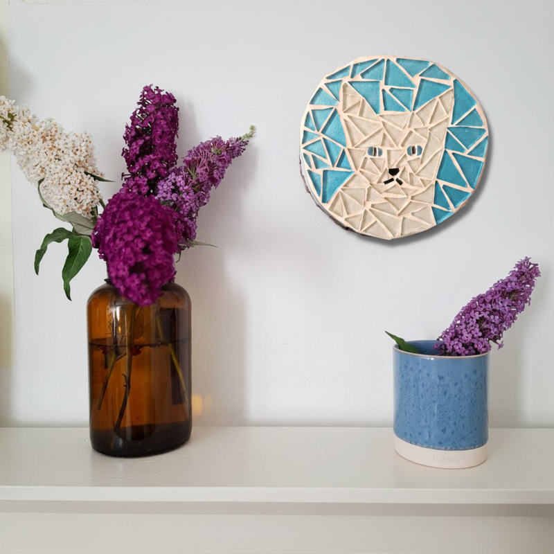 A circular mosaic wall art piece featuring a stylized cat face in white and turquoise, displayed on a white wall above a shelf with purple flowers in vases.