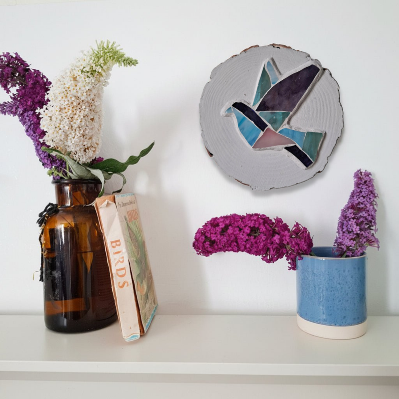 A styled interior shelf scene with flowers in an amber glass vase, a blue ceramic pot with purple flowers, and a round dove mosaic on a white wall.
