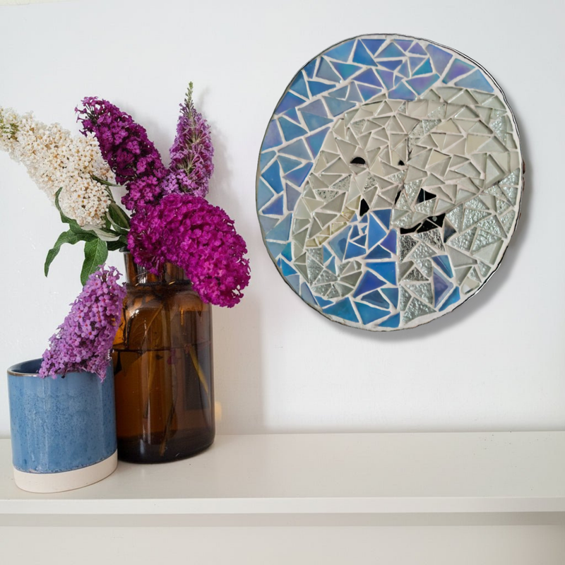 A round stained glass wall art piece featuring an elephant, displayed on a white wall above a shelf with purple and white flowers in vases.