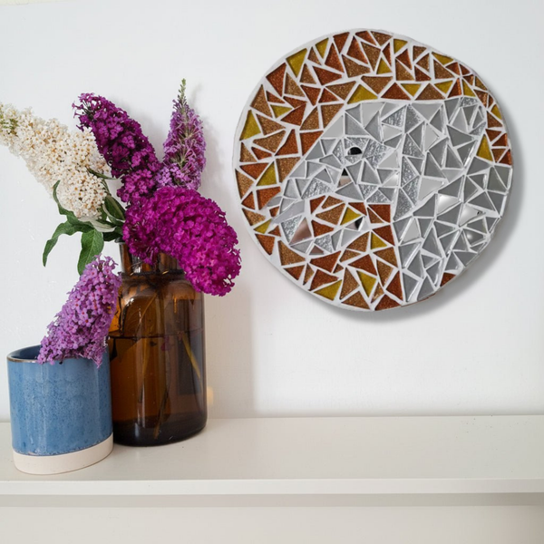 A round mosaic wall art piece featuring a stylized elephant face, displayed on a white wall above a shelf with decorative vases and flowers.