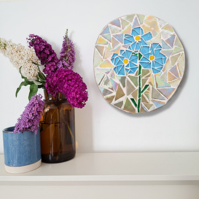 A round mosaic wall art piece featuring blue flowers with green stems, displayed on a white wall above a shelf with purple and white flowers in vases.