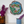 A round mosaic wall art piece featuring a frog design in green, blue, white, and brown tones, mounted on a white wall above a shelf. The shelf holds an amber glass vase with purple flowers on the left and a speckled blue ceramic pot with a purple flower on the right.