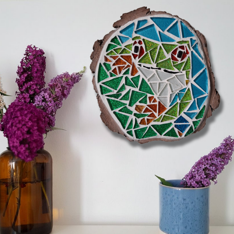 A round mosaic wall art piece featuring a frog design in green, blue, white, and brown tones, mounted on a white wall above a shelf. The shelf holds an amber glass vase with purple flowers on the left and a speckled blue ceramic pot with a purple flower on the right.