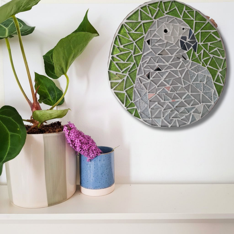 A circular mosaic wall art piece featuring a grey parrot on a green background, displayed on a white wall above a shelf with potted plants.