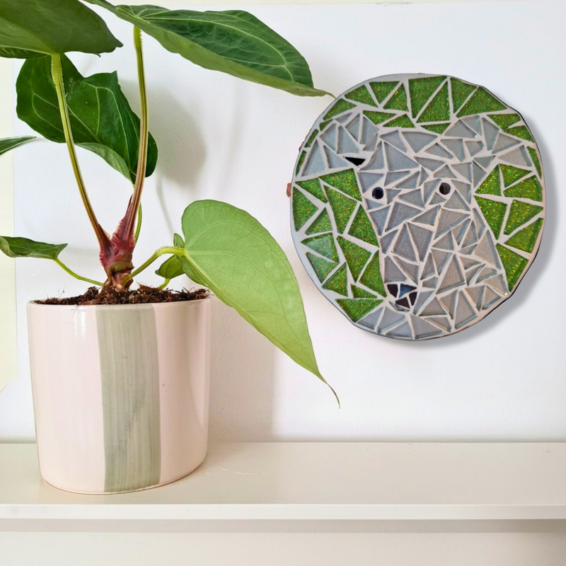 A round mosaic wall art piece featuring a greyhound made of green, grey, and white geometric pieces, displayed on a white wall next to a potted plant on a shelf.