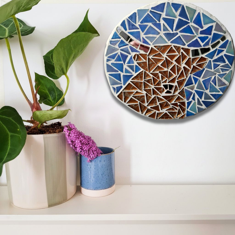 A round mosaic wall art piece featuring a stylized highland cow in shades of blue, brown, and white, mounted on a white wall above a shelf with potted plants.