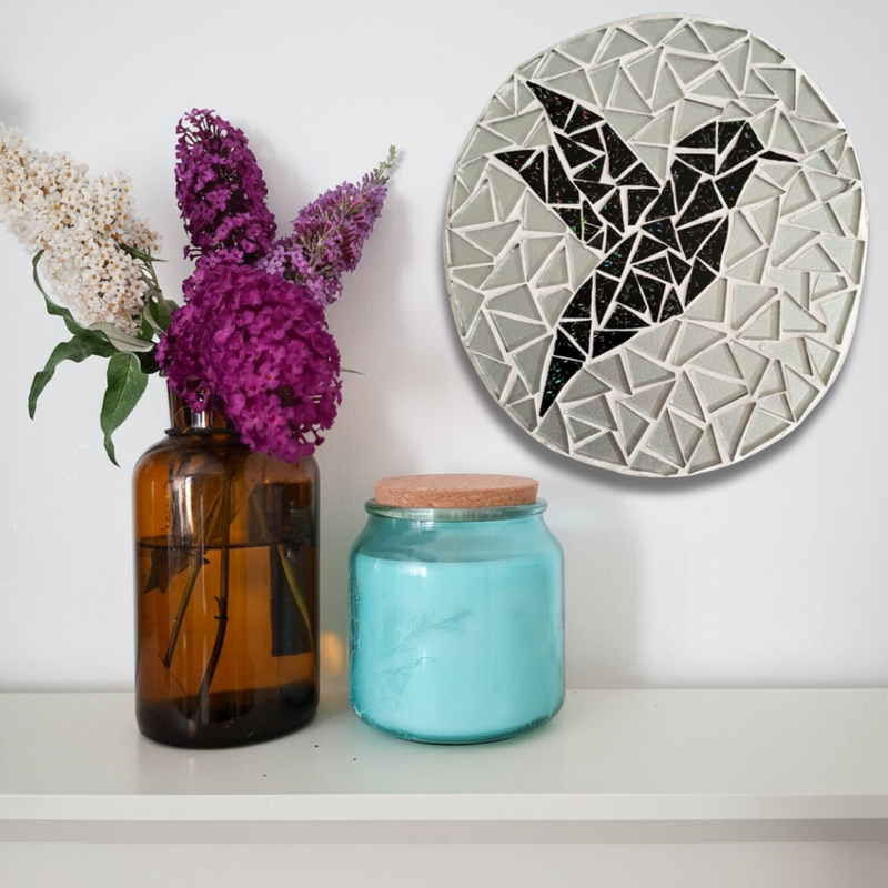 A round mosaic wall mirror with a black bird design hangs on a white wall above a shelf holding an amber glass vase with purple and white flowers and a turquoise glass jar with a cork lid.