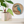 A round mosaic wall art piece featuring a kingfisher bird in blue, orange, and brown tones, mounted on a white wall beside a potted green plant on a white shelf.