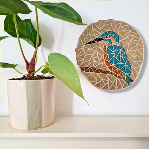 A round mosaic wall art piece featuring a kingfisher bird in blue, orange, and brown tones, mounted on a white wall beside a potted green plant on a white shelf.