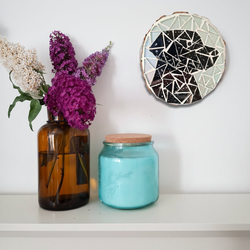 A styled interior scene showing a round mosaic of a black labrador hanging on a white wall above a shelf. On the shelf are an amber glass vase with white and purple flowers and a turquoise glass jar with a cork lid.