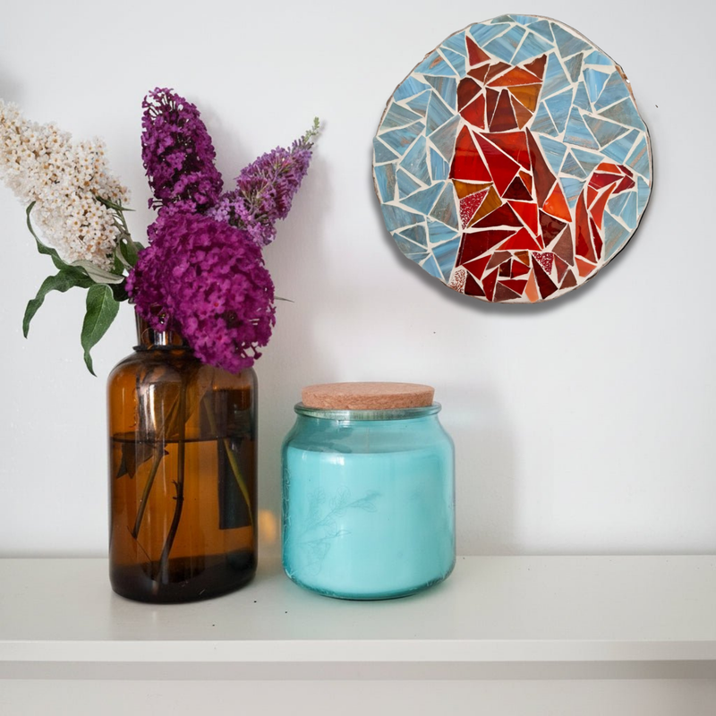 A styled interior shelf scene with a mosaic-style wall art piece featuring a red and orange cat on a blue and beige geometric background, hanging above an amber glass vase with purple and white flowers and a turquoise glass jar with a cork lid.