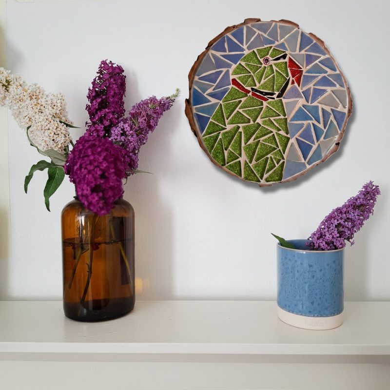 A styled interior scene showing a round mosaic wall hanging with a green bird design above a shelf. The arrangement includes an amber glass vase with white and purple flowers on the left and a blue ceramic pot with purple flowers on the right against a white wall.