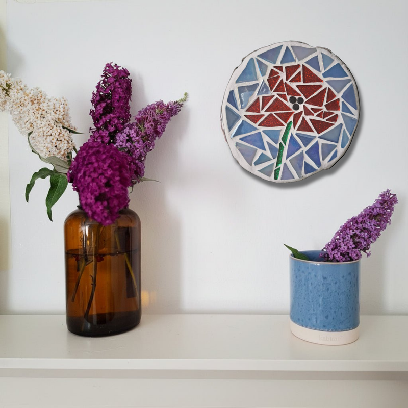 A styled interior scene with purple flowers arranged in two vases on a white shelf against a white wall: an amber glass bottle on the left and a speckled blue ceramic cup on the right. A round mosaic of a poppy is also visible above the shelf.