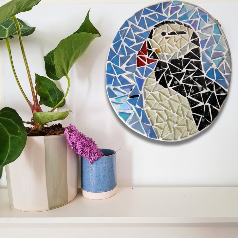 A circular mosaic wall art piece featuring a puffin in blue, black, white, and red, displayed on a white wall above a shelf with potted plants.