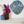 A round mosaic wall art piece featuring snowdrops hangs on a white wall above a shelf with two vases of flowers.
