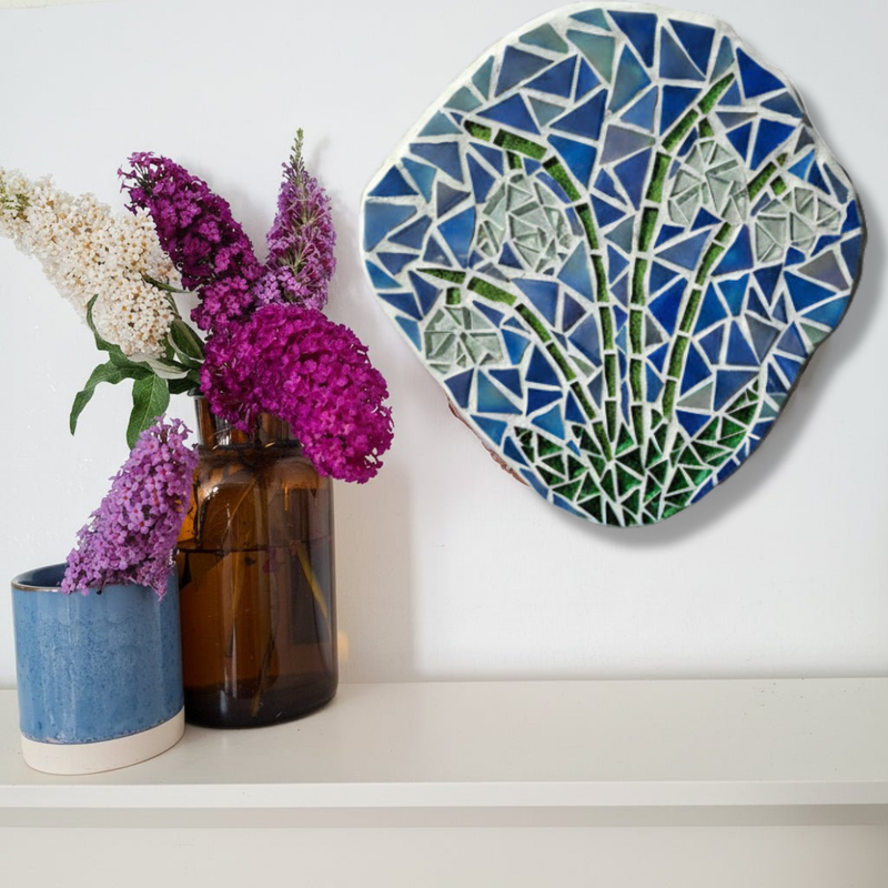 A round mosaic wall art piece featuring snowdrops hangs on a white wall above a shelf with two vases of flowers.