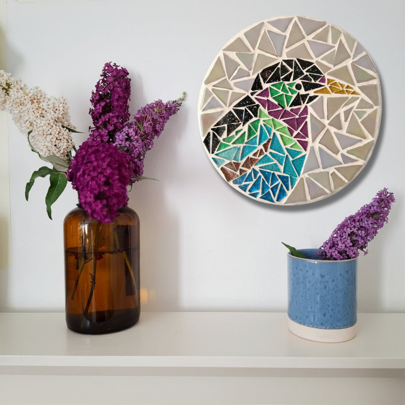 A circular mosaic wall art piece featuring a stylized bird in geometric segments, displayed on a white shelf next to two flower vases against a white wall.