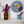 A round mosaic wall art piece featuring a toucan in shades of blue, yellow, black, and orange, displayed on a white wall above a shelf with purple flowers in vases.