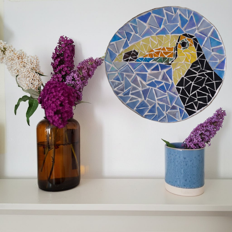 A round mosaic wall art piece featuring a toucan in shades of blue, yellow, black, and orange, displayed on a white wall above a shelf with purple flowers in vases.